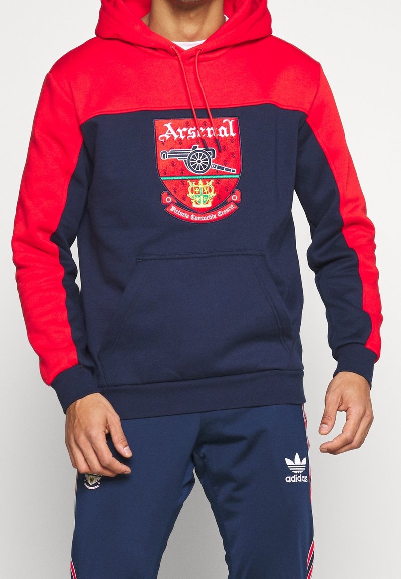 Huvtröja med marinblå kropp och röda ärmar. Har en Arsenal FC-logo på framsidan, kenguruficka och ribbade muddar.