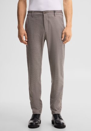 Mężczyzna noszący jasnobrązowe teksturowane spodnie i czarne sznurowane eleganckie buty, stojący na jednolitym tle.