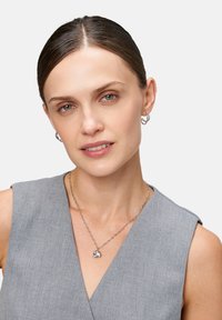 Silberner Elefanten-Anhänger an einer zarten Kette, kombiniert mit kleinen silbernen Creolen. Das Model trägt ein graues ärmelloses Oberteil.