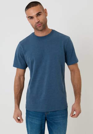 Uomo con capelli corti che indossa una maglietta blu a girocollo e jeans blu, in piedi davanti a uno sfondo chiaro e uniforme.