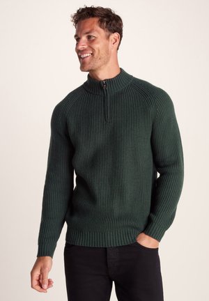 Dark green knitted sweater with ribbed texture, zip neck, and fitted cuffs. Worn over black pants, showcasing a snug design.