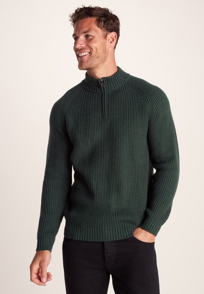 Dark green knitted sweater with ribbed texture, zip neck, and fitted cuffs. Worn over black pants, showcasing a snug design.