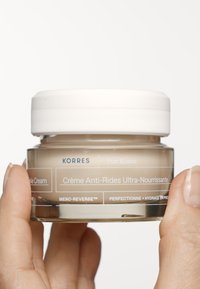 Korres anti-aging kräm i en klar glasbehållare. Rund form med ett vitt lock, prytt med blå och vita textetiketter för produktinformation.
