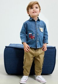 Camicia di denim con ricamo di un trattore rosso sulla tasca, pantaloni cargo color khaki e scarpe bianche con cinturini in velcro, posizionati su una panchina blu.