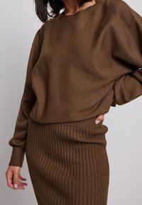 Maglione marrone lavorato a maglia con una vestibilità rilassata, scollatura rotonda e polsini a costine, abbinato a una gonna marrone a costine, creando un outfit coordinato.