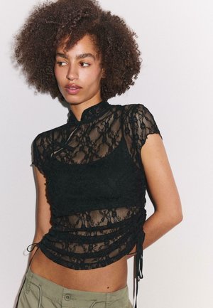 Mladá žena s kučeravými vlasmi sa pozerá do strany, má na sebe čierny priehľadný čipkovaný top cez čierny bralette a zelené nohavice na bielom pozadí.