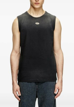 Homme portant un débardeur noir délavé sans manches avec un petit logo sur la poitrine et un jean bleu foncé, debout les bras détendus le long du corps.