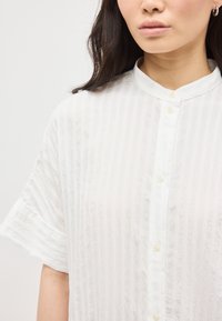 Γυναίκα με μεσαίου μήκους σκούρα μαλλιά, φορώντας ένα λευκό πουκάμισο με κοντά μανίκια και λεπτές κατακόρυφες ρίγες, καθώς και ασημένια σκουλαρίκια κρίκους.