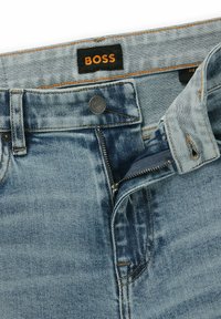 Vaaleansiniset farkut, joissa on vetoketjulahke ja nappisulkimet. Ominaisuuksia ovat tummanruskea "BOSS" -tunnus ja oranssitompeluseikat.