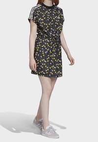 Krátké černé šaty s květinovým vzorem, elastickým pasem, kulatým výstřihem a krátkými rukávy. Jsou doplněny bílými pruhy na ramenech a světlejšími teniskami.