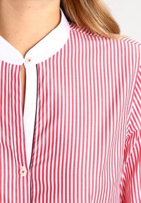 Blusa a righe rosse e bianche con colletto alto. Presenta una chiusura a bottoni e un tessuto testurizzato. Nota per le sue righe verticali.