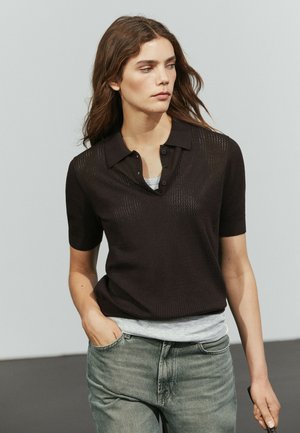 Giovane donna con lunghi capelli castani indossa un top nero a maniche corte sopra una camicia grigia e jeans verdi sbiaditi, con una mano in tasca.