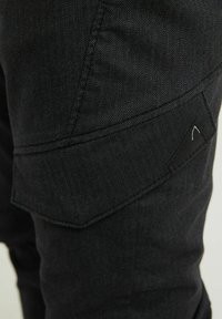 Close-up van zwarte gestructureerde stoffen broek met schuin flapzakje en zichtbare stikseldetails.