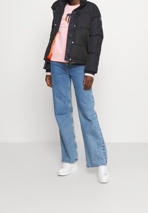 Osoba ubrana w czarną kurtkę puchową na różowej bluzie, jasnoniebieskie szerokie dżinsy i białe sneakersy na platformie, stojąca na jednolitym tle.