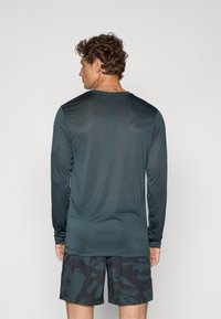 Langærmet teal træningsskjorte lavet af glat, letvægtsstof, med rund halsudskæring og subtil tekstur, parret med mørke mønstrede shorts.