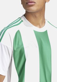 Grüne und weiße Trikot mit drei grünen Streifen auf den Schultern, aus strukturiertem Stoff. Verfügt über ein geprägtes Adidas-Logo auf der Brust.