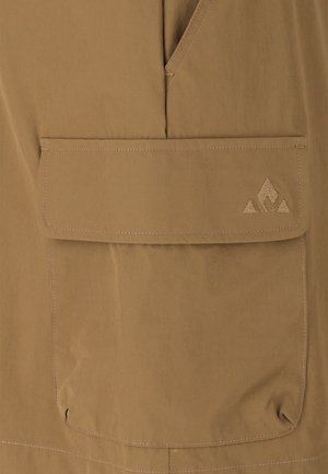 Lähikuva vaaleanruskeasta takin taskusta, jossa on läppä ja ommeltu logo, jossa kolme kolmion muotoista elementtiä alaoikeassa kulmassa.