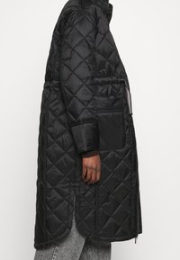 Abrigo largo acolchado negro con cuello alto, bolsillos laterales y un acabado suave y reflectante. Presenta un corte recto y una textura suave.