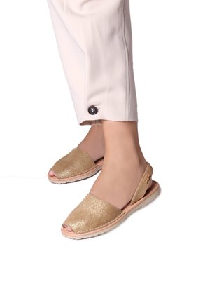 Sandales à talon slingback dorées pailletées avec orteils ouverts, semelles plates et un accent beige à l'arrière. Portées avec un pantalon clair à bouton.