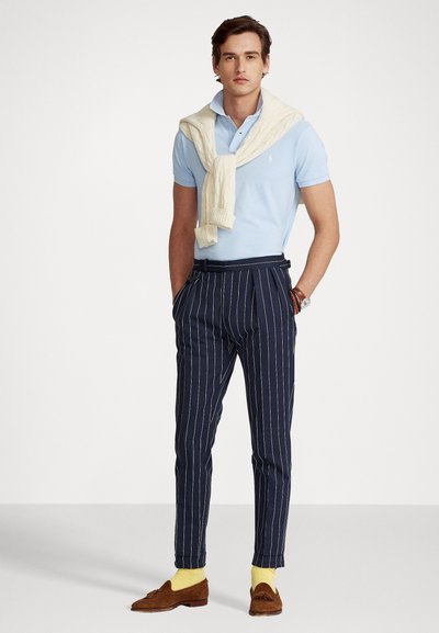 Light blue polo shirt, cream knitted sweater draped over shoulders, navy pinstriped trousers, brown suede loafers, and yellow socks.