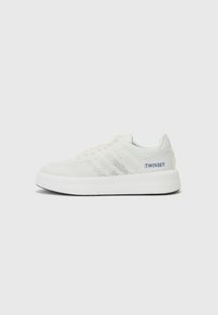 Hvit sneakers med mesh-overdel, teksturdetaljer, rund tå og kontrasterende marineblå logo. Har en tykk, hvit gummisåle.