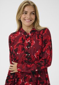 Rochie roșie cu flori, având modele de flori alb-negru, guler la gât și accente din perlă pe butoanele din față. Textură de țesătură netedă.