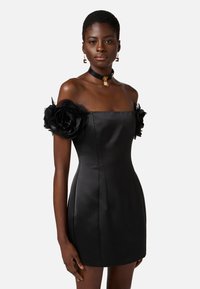Abito off-the-shoulder in raso nero con grandi rose di tessuto nero sulle maniche, corpetto aderente e una mini gonna dritta. Collana elegante e orecchini.
