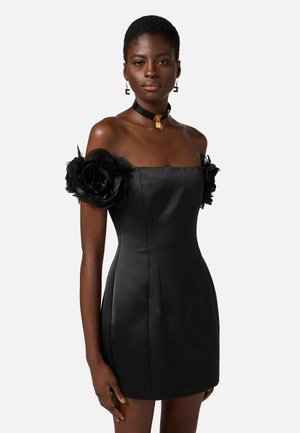 Abito off-the-shoulder in raso nero con grandi rose di tessuto nero sulle maniche, corpetto aderente e una mini gonna dritta. Collana elegante e orecchini.