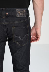 Mörka denimjeans med orange sömmar, som har en synlig logotyp på midjebandet och en distinkt design på bakfickan.