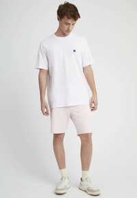 T-shirt en coton blanc avec un petit motif bleu sur la poitrine gauche, associé à un short rose clair. Porté avec des baskets beiges et blanches et des chaussettes jaunes.
