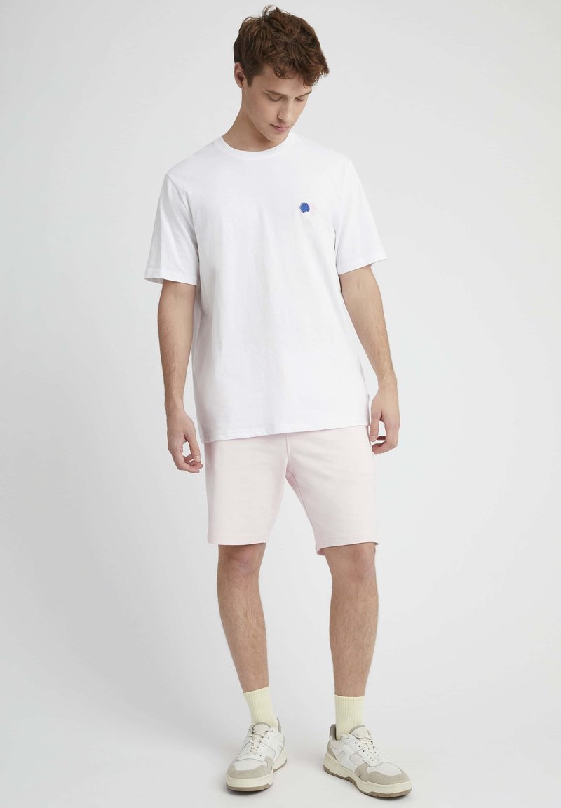 T-shirt en coton blanc avec un petit motif bleu sur la poitrine gauche, associé à un short rose clair. Porté avec des baskets beiges et blanches et des chaussettes jaunes.