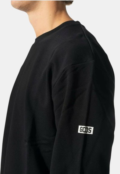 Mann trägt einen schwarzen Pullover mit einem weißen "GCDS"-Logoaufnäher am linken Ärmel, Hals und Kieferpartie vor einem schlichten Hintergrund sichtbar.