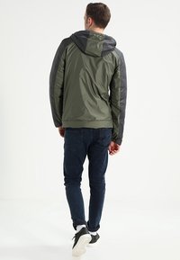 Olivgrön jacka med svarta ärmar, med huva och slät, blank yta. Bärs med mörka jeans och avslappnade skor.