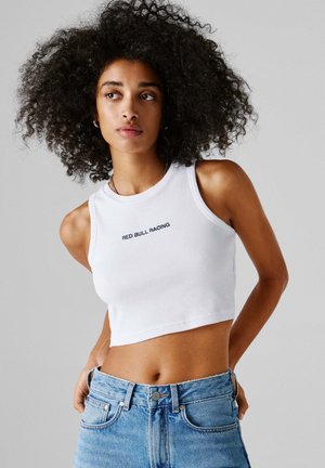 Mlada žena s kovrčavom kosom nosi bijelu kratku majicu s natpisom "RED BULL RACING" i plave traperice na sivoj pozadini.