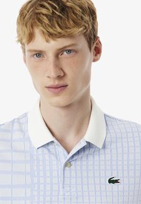 Ljusblå polo skjorta med rutigt mönster, vit krage och en grön krokodil-logotyp på bröstet. Texturen ser slät och avslappnad ut.