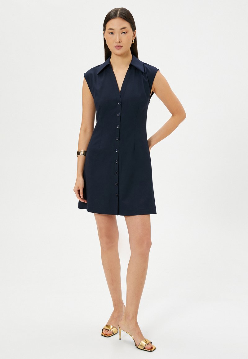 Robe courte marine avec un col aiguisé, des manches courtes, une fermeture à boutons sur le devant et une coupe ajustée. Portée avec des sandales à talons dorées.