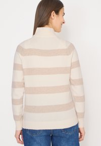 Femme de dos, portant un pull crème avec des rayures horizontales beige et un jean bleu, sur un fond blanc uni.