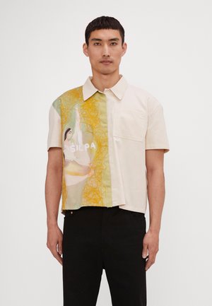 Short-sleeve shirt featuring a cream-colored fabric with an artistic green and yellow printed panel, including a graphic design and pocket.