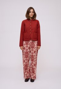 Giacca trapuntata rossa con chiusura a bottone e due tasche anteriori, abbinata a pantaloni panna a fantasia con disegni floreali e geometrici rossi.