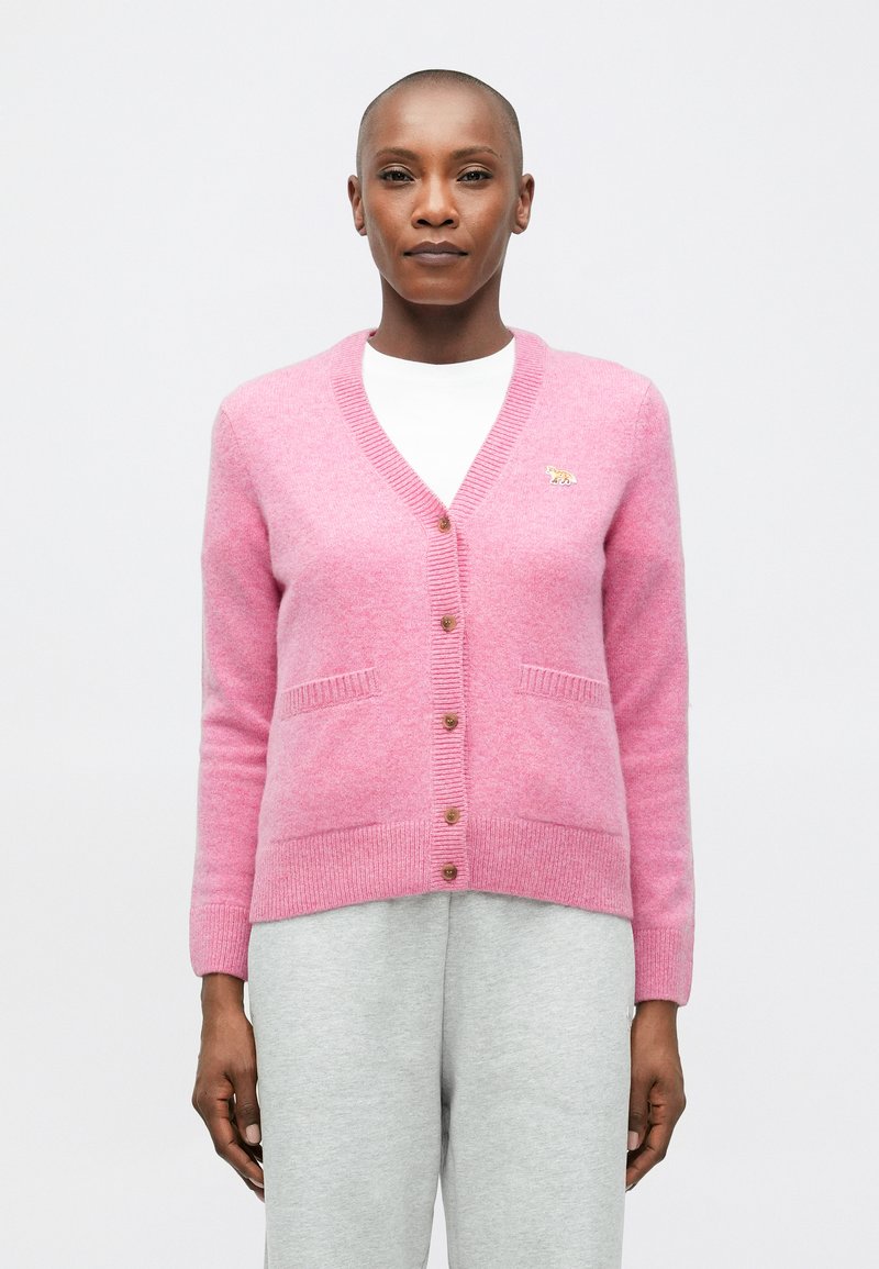 Cardigan roz tricotat, cu decolteu în V, închidere cu nasturi, buzunare și o mică accentuare aurie. Textură moale și detalii cusute pe margini.