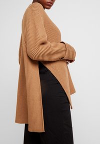 Lång, randig cardigan i kamelfärg, med V-ringning, sänkta axlar och asymmetrisk fåll. Buren över svarta byxor.