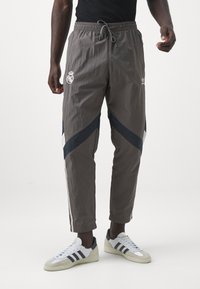 Pantalones deportivos grises con cintura elástica, con detalles en azul marino y franjas blancas, que muestran los logotipos de Real Madrid y Adidas. Combinados con zapatillas blancas.
