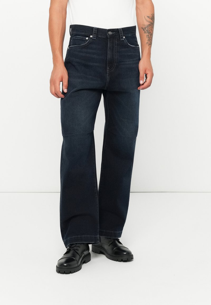Dark blue wide-leg denim jeans with five pockets, button closure, and subtle detailing; paired with black lace-up ankle boots.