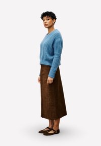Femme aux cheveux courts et bouclés portant un pull en tricot bleu, une jupe midi marron et des ballerines Marron Mary Jane, se tenant de profil.