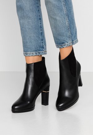 Czarne skórzane botki za kostkę z zamszowym panelem, okrągłym noskiem oraz słupkowym obcasem z złotym akcentem. Noszone z przyciętymi jasnoniebieskimi dżinsami.