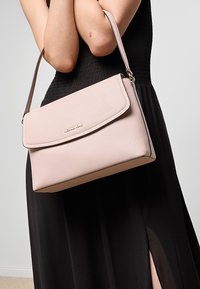 Una donna che indossa un vestito nero senza maniche tiene una borsa a tracolla in pelle rosa chiaro con patta e logo dorato.