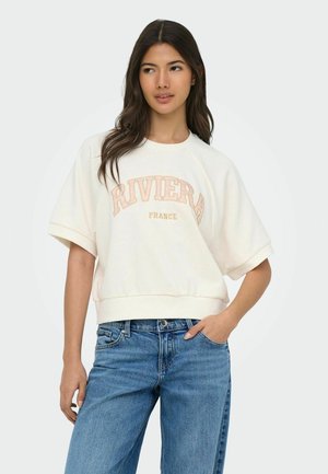 Młoda kobieta o długich ciemnych włosach, ubrana w kremową bluzę z krótkim rękawem z napisem "RIVIERA FRANCE" oraz niebieskie dżinsy, pozująca z jedną ręką w kieszeni.
