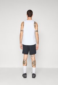 Haut sans manches blanc associé à un short noir. Le tissu semble léger, avec des tatouages visibles sur les bras et les jambes. Des chaussures de sport noires sont portées.