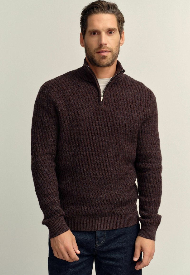 Strickpullover in Dunkelbraun mit einem Reißverschlusskragen. Verfügt über ein strukturiertes, geripptes Design und eng anliegende Bündchen, kombiniert mit einer dunklen Jeans.