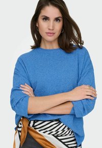 Femme aux cheveux bruns portant des boucles d'oreilles créoles en or et une bague, pull bleu, et écharpe rayée noire, blanche et brune nouée à la taille, bras croisés.